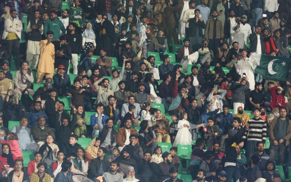 A large crowd of people eagerly watching a cricket event in the PSL tournament, seated in a stadium with green seats. Some are standing, taking photos, and waving flags. Small children are visible among the adults, and the atmosphere appears lively and engaged. | jeetwinbet.com
