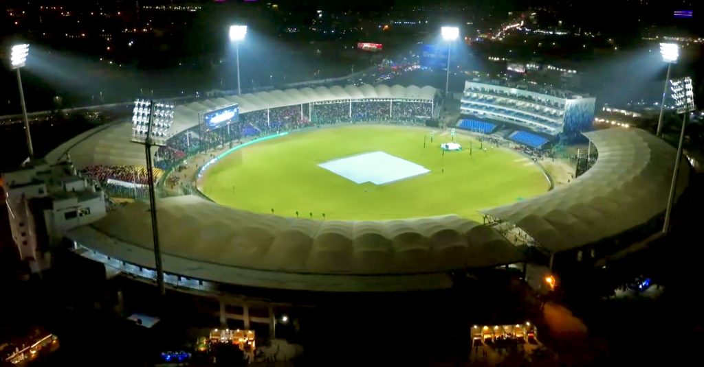 Aerial view of a brightly lit cricket stadium at night, with a green field and a covered white pitch in the center. The stadium is surrounded by seating areas filled with spectators, and city lights are visible in the background. | jeetwinbet.com
