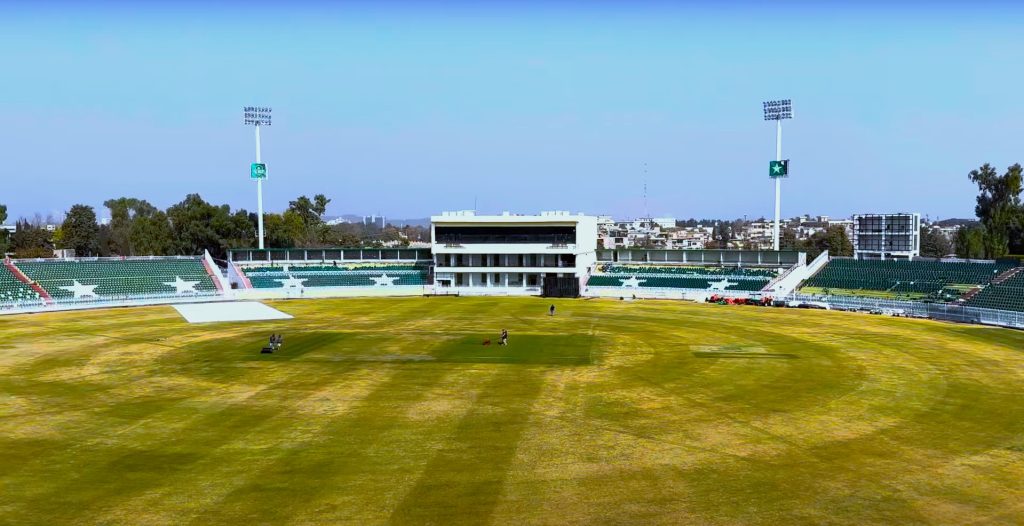 A cricket stadium featuring a large green field surrounded by tiered seating. The stands are partially filled with chairs, and a few people can be seen on the field surface. Tall floodlights are positioned at various points around the stadium. | jeetwinbet.com