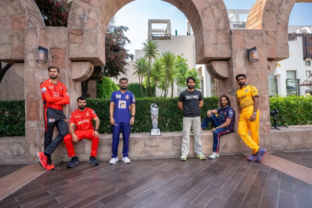 Six men stand and sit around a trophy under stone arches. They wear different colored sports uniforms, with one in casual attire. The setting includes greenery and a modern building in the background. | jeetwinbet.com Pakistan Super League Preview: Teams & Format