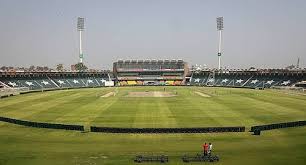 A wide-angle view of a cricket stadium with green grass and an empty pitch in the center. Tall floodlights stand at the corners. The stands are empty, and a few people can be seen near the boundary. | jeetwinbet.com