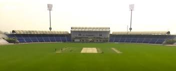 A cricket stadium with empty stands. The field is green, featuring a cricket pitch in the center. There are floodlights towering over the seating areas, and a large building is visible in the background. | jeetwinbet.com