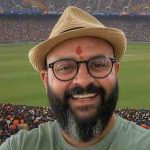 A man with a beard, glasses, and a beige hat smiles at the camera. He has a red mark on his forehead. A large crowd fills the stadium in the background, with a green field visible. | jeetwinbet.com